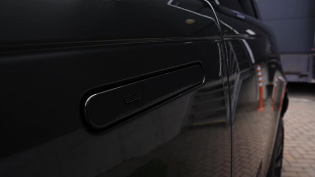 Low angle close up shows glossy pop out handle with button on black SUV. Reflections show brick paving, trailer, and orange bollards in an industrial area.