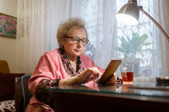 Elderly woman using smart phone under electric lamp on table in living room