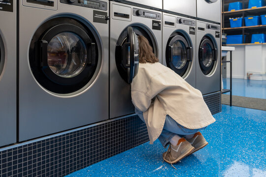 Young woman squatting and looking inside of washing machine at laundromat