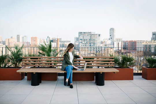 Woman using laptop while sitting on wooden bench at building terrace