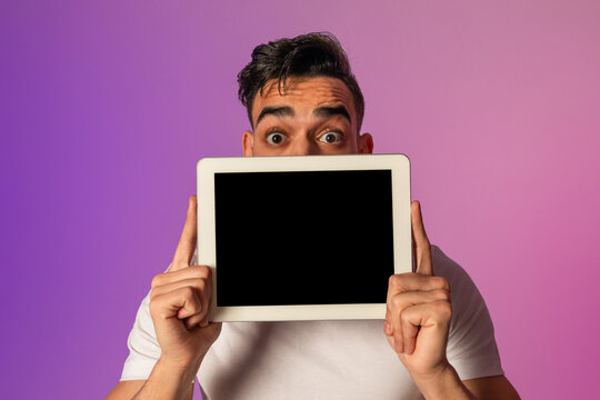 A surprised millennial Arab man hides behind a tablet with a blank screen while illuminated by neon lights. He offers space for advertisements, showcasing a modern digital approach.