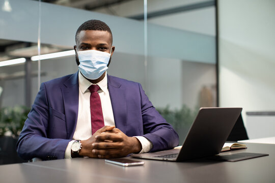 Portrait of african american young man in suit manager in face mask posing while working in office, sitting at worktable in front of laptop, looking at camera. Business while coronavirus epidemic - Powered by Adobe
