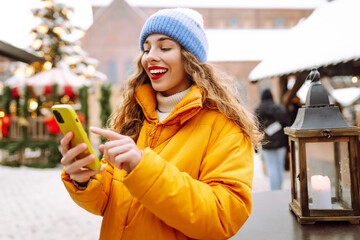 Portrait of woman using phone, enjoying Christmas atmosphere at market. Beautiful woman takes...