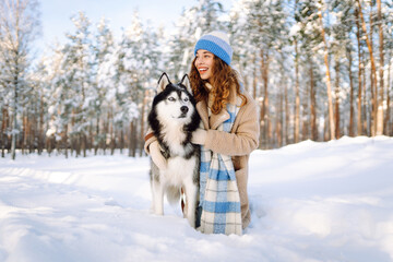 A cheerful woman in a warm coat and blue hat walks her husky in a snowy forest. A beautiful woman and her pet enjoy a winter day. Concept of friendship and fun.
