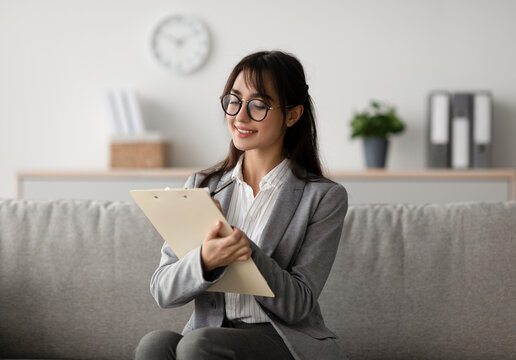 A focused young arab female psychologist sits on a couch in a modern office, writing in a clipboard while offering professional counseling services to her clients.