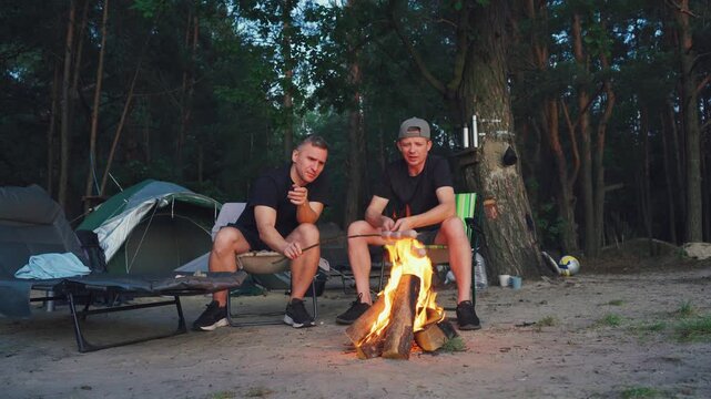 Laughter and stories beside glowing campfire, men enjoying friendship and stories in tranquil woodland scene, relaxed group sharing humor and tales as night falls in peaceful forest setting