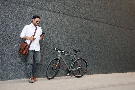 Man leaning on wall checking phone near bicycle