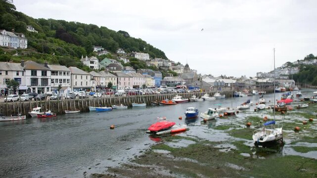 Boats anchored in the harbor at low tide in Looe, Cornwall, England, with colorful houses and a church tower visible on the tree-covered hill. Mooring buoys and seaweed line the water&rsquo;s edge.