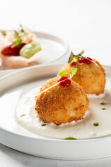 A close-up shot of two golden-fried arancini or croquettes served with creamy sauce on a white plate, garnished with microgreens and red dots