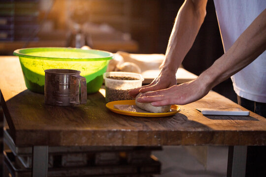 Hands kneading dough on yellow plate, preparing bread or pastry on wooden table with green bowl, metal sifter, and container of grains, warm light illuminating baking process - Powered by Adobe