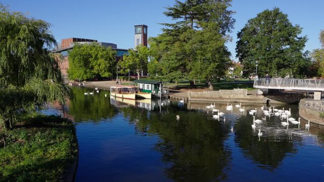 A static wide angle view of Cox&rsquo;s Island and tour boats moored on the River Avon, framed by trees with the modern Royal Shakespeare Theatre and tower in the background in Stratford-upon-Avon.