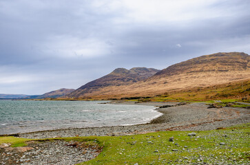 Peaceful bay with waves, stones, and rolling hills on the Isle of Mull, Scotland. Natural coastal scenery with soft light and cloudy weather.