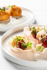 A vibrant close-up of modern fine-dining appetizers on white plates. Foreground: prawn crackers with tuna poke, avocado, and greens. Background: crispy fried arancini