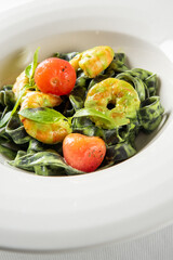 A close-up vertical studio shot of vibrant green spinach tagliatelle pasta with prawns, cherry tomatoes, and basil leaves in a white bowl.