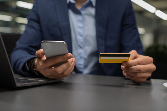 Entrepreneur in a suit sits at an office desk, using a smartphone in one hand and holding a credit card in the other. He is shopping online while working on a laptop in a modern workspace. - Powered by Adobe