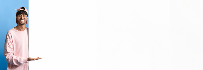 A cheerful man wearing a pink sweater and a cap stands against a blue background. He is smiling and gesturing as if inviting viewers to pay attention.