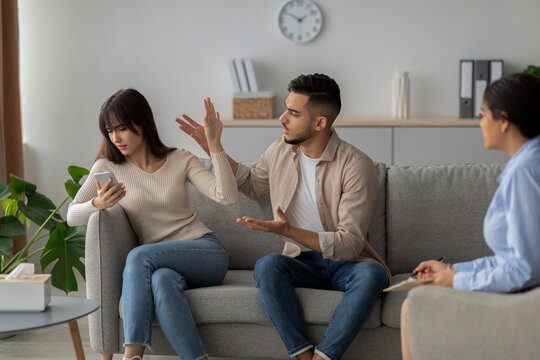 An angry couple discusses their relationship issues with a therapist. The man gestures expressively while the woman appears focused on her phone.