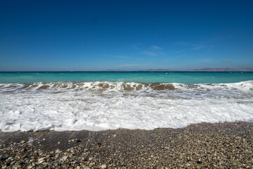 Turquoise sea wave with white foam washing over pebble beach under clear blue sky and distant islands on horizon.