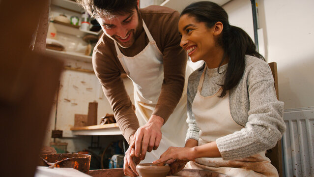 Male Teacher Helping Woman Sitting At Wheel In Pottery Class