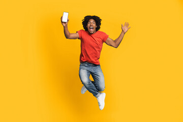 A young man dressed in a red shirt and jeans leaps joyfully in the air, holding up a smartphone. His wide smile and energetic pose convey a sense of excitement and celebration.