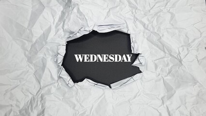 Image featuring the word "WEDNESDAY" visible through a torn hole in heavily crumpled white paper, revealing a dark background.