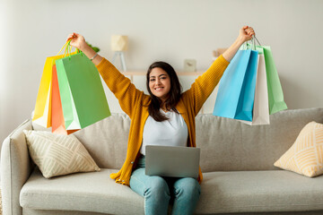 An excited Arab woman sits on her sofa with a laptop, raising colorful gift bags above her head in happiness. She enjoys shopping online and is pleased with her purchases and discounts.