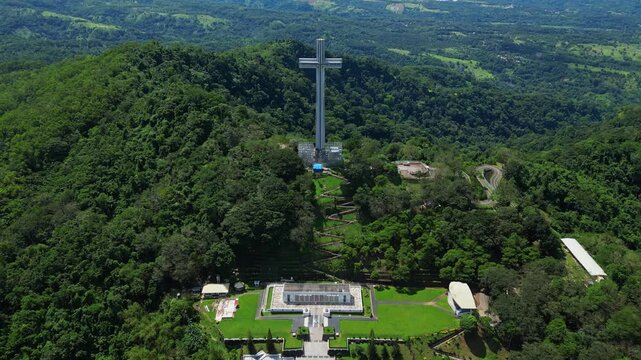 Front‑view push aerial highlighting the towering Mt. Samat National Shrine, revealing its monumental cross framed by landscaped grounds and lush forested hills in Pilar, Bataan, Philippines.
