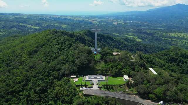 Wide pull‑out aerial revealing the monumental Mt. Samat National Shrine, showcase its towering cross framed by lush forests, valleys, and distant mountains in Pilar, Bataan, Philippines.