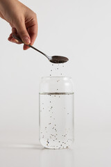 Pouring chia seeds from a silver spoon into a glass of water on a white background
