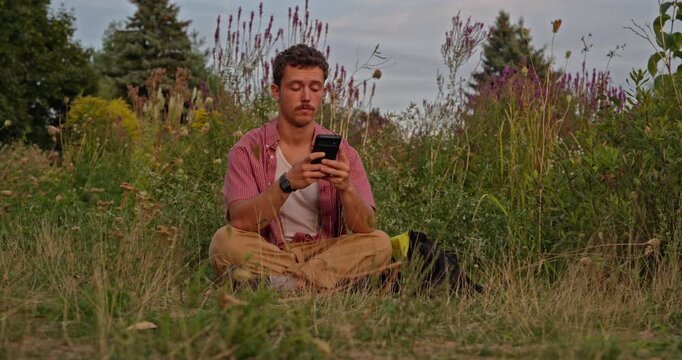 A young Gen Z man sits in a wild meadow, checks his phone, becomes tired, then disconnects to appreciate the cloudy colorful sky and meditate in nature. Perfect for digital detox themes.