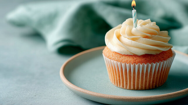 A vanilla cupcake with glossy cream frosting and a lit striped candle sits on a soft green plate, surrounded by gentle fabric creating a calm, inviting atmosphere.