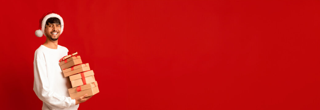 A young man smiles while carrying several neatly packaged gift boxes tied with red ribbons. He wears a cozy white sweater and a Santa hat, celebrating the holiday spirit in a vibrant setting. - Powered by Adobe