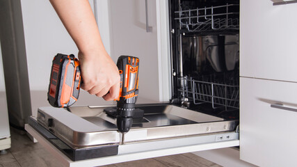The hands of a craftsman with a screwdriver install a kitchen front panel on a dishwasher, industry