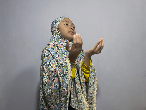 Little Muslim girl dressed in a floral-patterned mukena praying with raised hands in devotion to Allah.