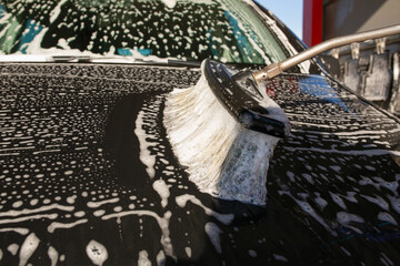 Detailed close up of a large foam covered brush washing the hood of a black car, showing soap streaks, bubbles and the cleaning process during an outdoor car wash.