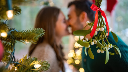 Close-up of festive mistletoe with a red ribbon in soft holiday lights, while a couple shares a warm, blurry kiss in the background, creating a cozy and romantic Christmas atmosphere