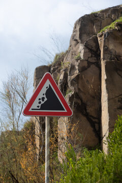 A falling-rocks warning sign stands against a rocky cliffside, alerting drivers to possible landslides in the area. The triangular red-bordered sign contrasts with the natural landscape of stone