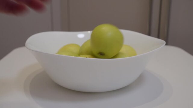 A fresh apple is dropped into a ceramic bowl already containing other apples. The apple impacts the others and the bowl, creating a light thump and clunk.