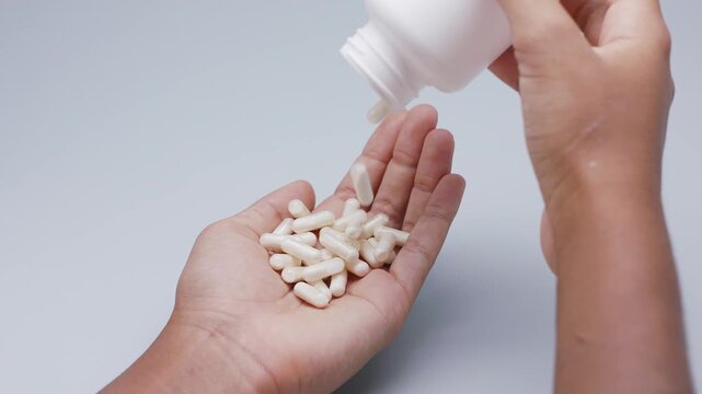 White capsules or pills being poured from a plastic bottle into a hand against a light gray background. Concept of medicine, healthcare, supplements, and pharmaceutical treatment.
