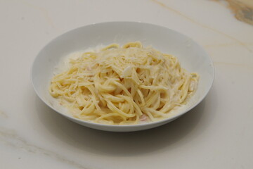 a white plate of spaghetti with carbonara sauce, with ham and grated cheese on a white veined marble table