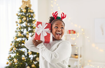 Portrait of joyful African American girl standing in beautifully decorated room, holding red gift...