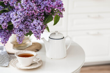 The concept of home comfort.A large purple bouquet of lilacs, white Scandinavian houses, a ceramic teapot and a cup on the table. In the background is the interior of a modern white kitchen.