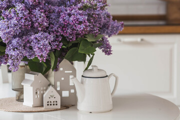 The concept of home comfort. A large purple bouquet of lilacs, white Scandinavian houses and a ceramic teapot on the table. In the background is the interior of a modern white kitchen.