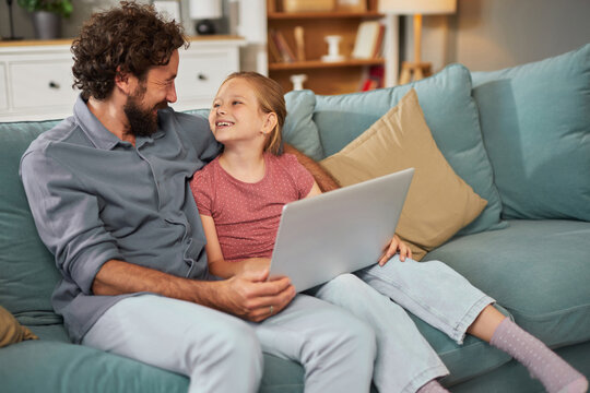 A father and his young daughter share smiles and laughter while sitting on a comfortable couch. They engage with a laptop, creating a warm family bonding moment in their home.