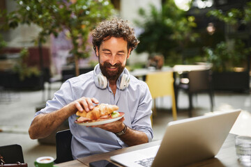 A man with curly hair and headphones sits at a table, smiling as he prepares to eat a croissant while working on his laptop in a lively outdoor cafe.