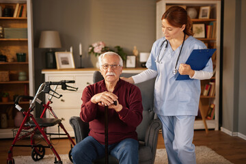 During a daytime home visit, a compassionate healthcare nurse gently puts her hand on the shoulder of an elderly man sitting in his armchair, with his cane, in a cozy living room, walker nearby.