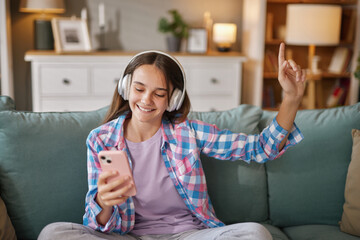 A teenage girl sits on a couch at home, wearing headphones and smiling while listening to music on her phone. She is relaxed and enjoying her leisure time in a cozy living room.