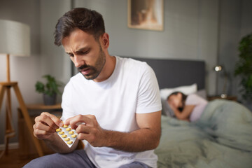 A man is focused on a strip of vitamins as his partner sleeps in the background, indicating a...