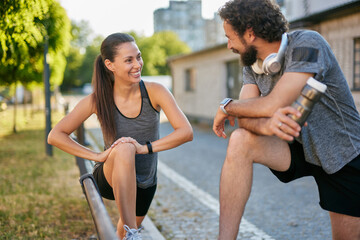Two individuals engage in a friendly stretching session in a park. They smile and chat, emphasizing camaraderie and a commitment to maintaining a healthy lifestyle outdoors.