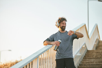 A man with headphones stands on a staircase by the river, checking his fitness watch while holding a water bottle. He looks relaxed and focused on his workout routine during the early morning. © Stockphotodirectors